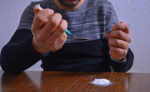 man putting meth from spoon into a syringe to inject it with white powder on table below him